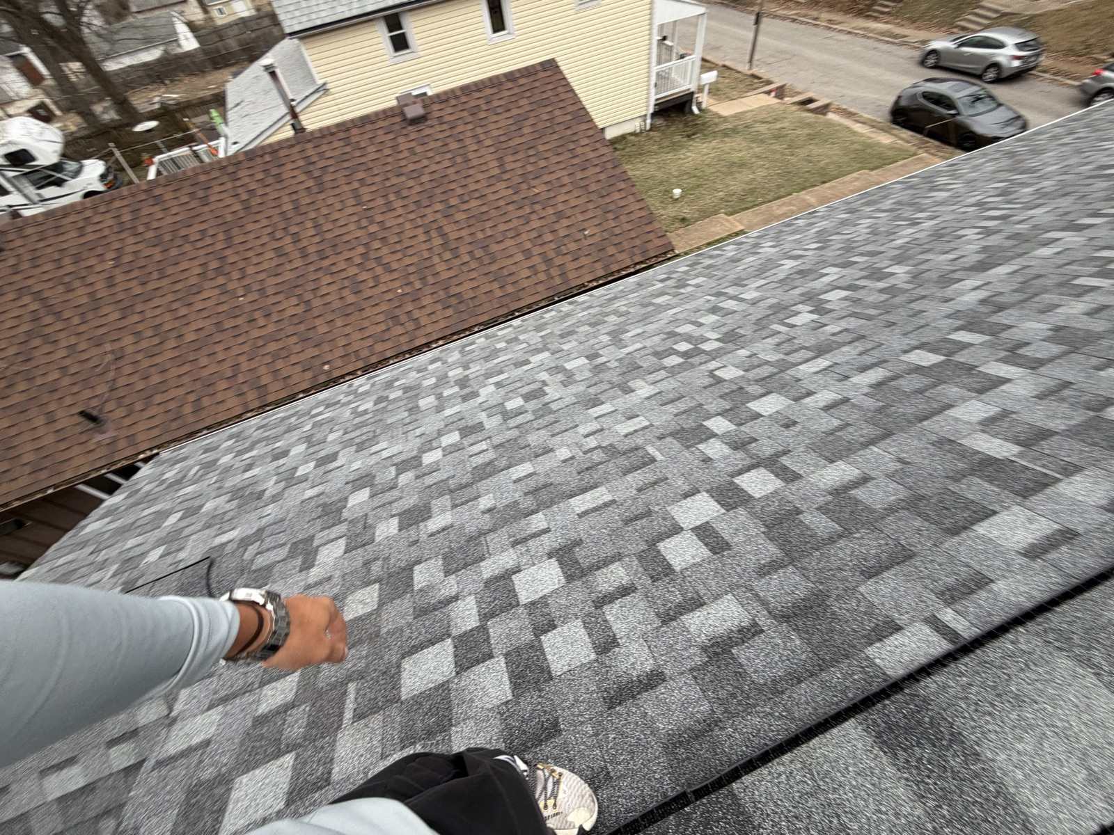 Downward view across a newly laid grey and charcoal architectural shingle roof taken by a person standing on it, their forearm with a metal wristwatch reaching into the frame at the left and their knee and trainer at the bottom. The adjoining lower roof along the left is still covered in older brown three tab shingles with their small even rectangular tabs, and beyond are a yellow vinyl sided house, a winter lawn and a street with parked cars.