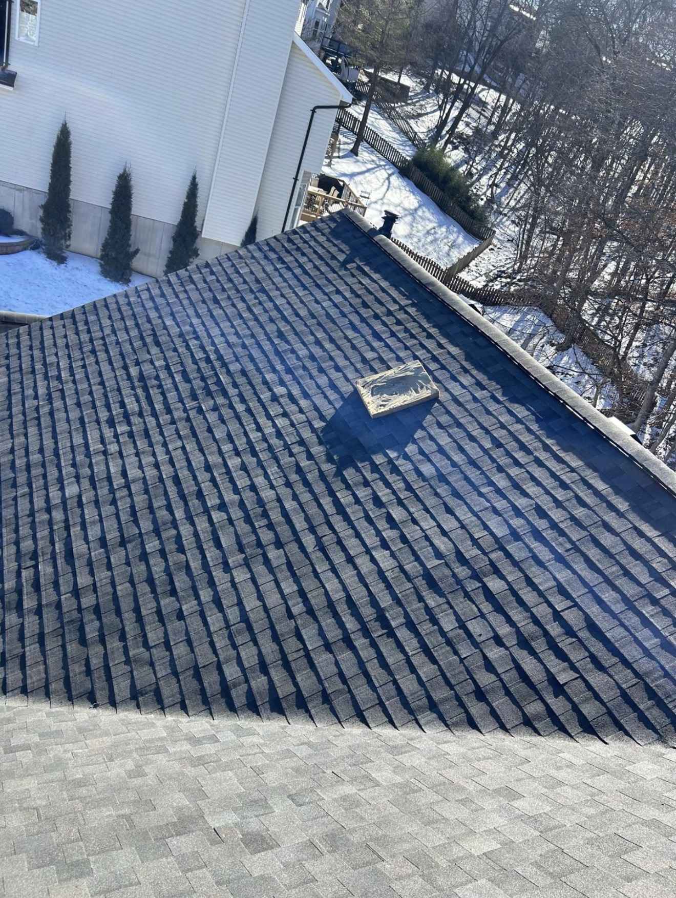 A large charcoal shingle roof plane part way through installation, courses running away up the slope with a hip line crossing diagonally and a lighter grey shingle surface visible along the lower edge of the frame. An opened wrapped bundle of shingles sits on the slope near a small vent, and beyond the roof edge are a white vinyl sided house wall with a downspout, clipped conifers, a wooden fence, lying snow and bare trees in hard low sun.