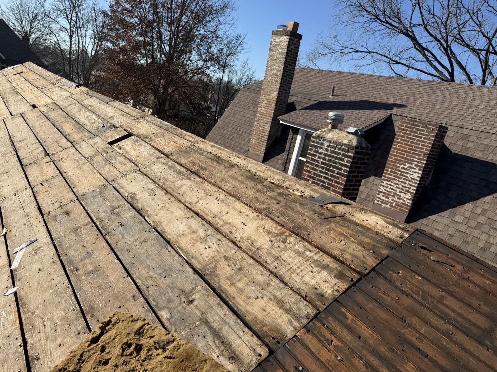A roof plane stripped to its original wide wooden plank decking running up the left of the frame, boards weathered grey and studded with old nails, with visible gaps between several boards and one board split away. Beyond it stands the rest of the house still carrying its weathered brown shingles, two brick chimneys with spalling faces and patched mortar, a small dormer with a metal turbine vent, and bare winter trees against a clear blue sky.