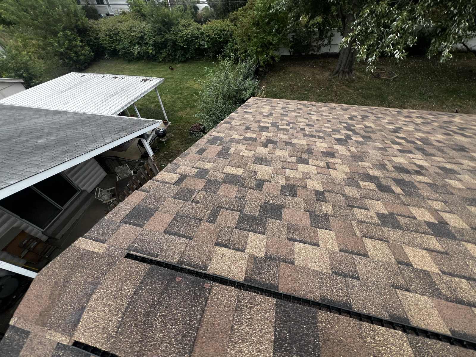 Wide overhead view from the ridge of a newly laid brown, tan and charcoal multi tone architectural shingle roof filling the right two thirds of the frame, with a separate lower grey shingled porch roof and a white corrugated carport canopy at the left, and a back garden below with a kettle barbecue, patio chairs, a hedge line and a large tree.