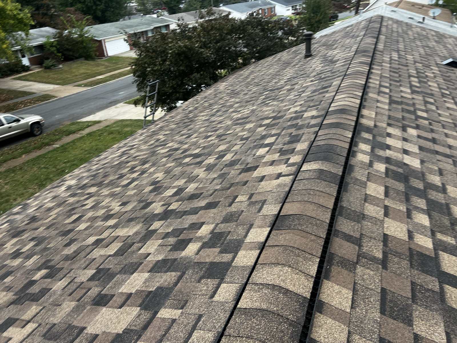 The capped ridge of a newly laid brown, tan and charcoal multi tone architectural shingle roof running diagonally away from the camera with a dark ridge vent beneath the cap, a metal flue pipe standing near the far end, and an aluminium ladder hooked over the eave at the left. Below is a suburban street of single storey brick and vinyl ranch houses with a white pickup parked at the kerb and mature trees along the verge.